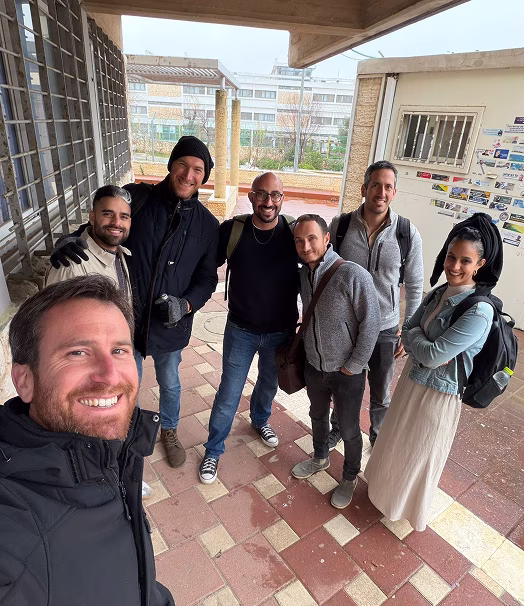 A group of seven people smiling together for a selfie outside an institutional building.