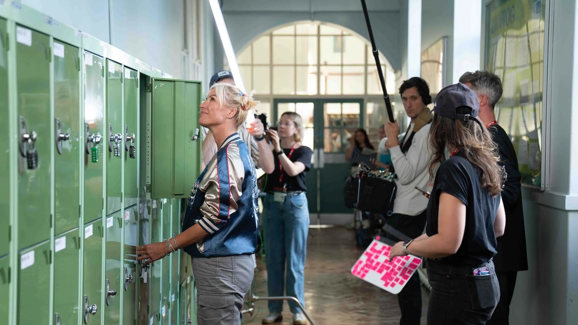 A woman in a blue satin jacket opens a green locker in a hallway while a film crew records her.