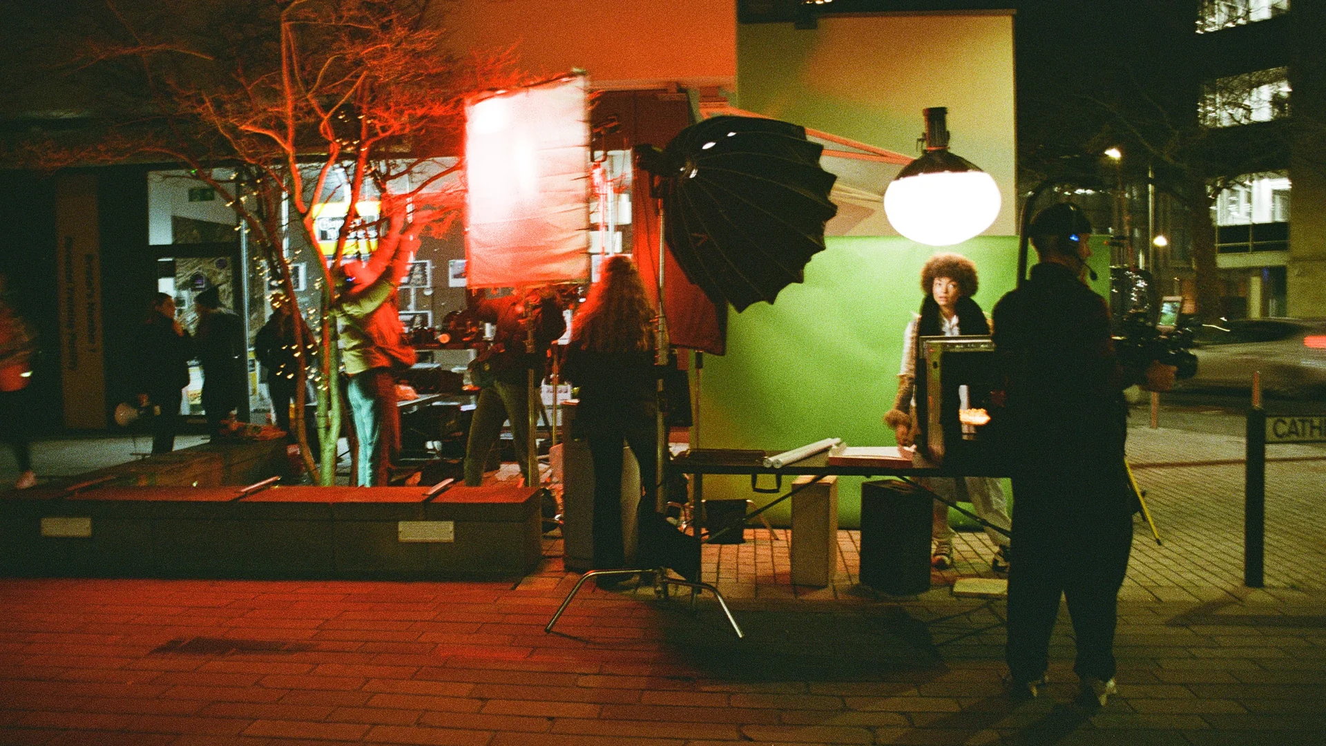 Nighttime outdoor film set with crew adjusting lighting and camera equipment around a person standing in front of a green screen.