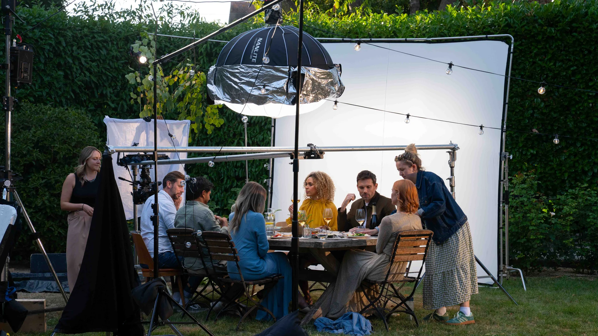 Film crew setting up outdoor dining scene with seven actors around a table with lighting and reflector equipment.