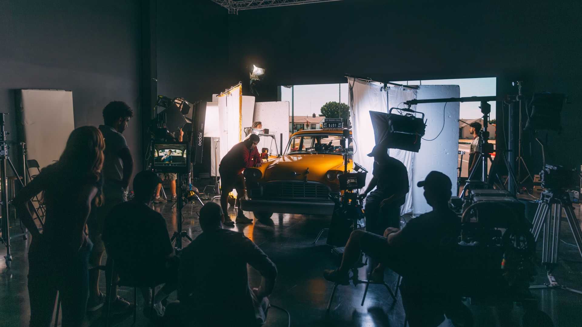Film crew setting up lighting and cameras around a vintage yellow car inside a studio.