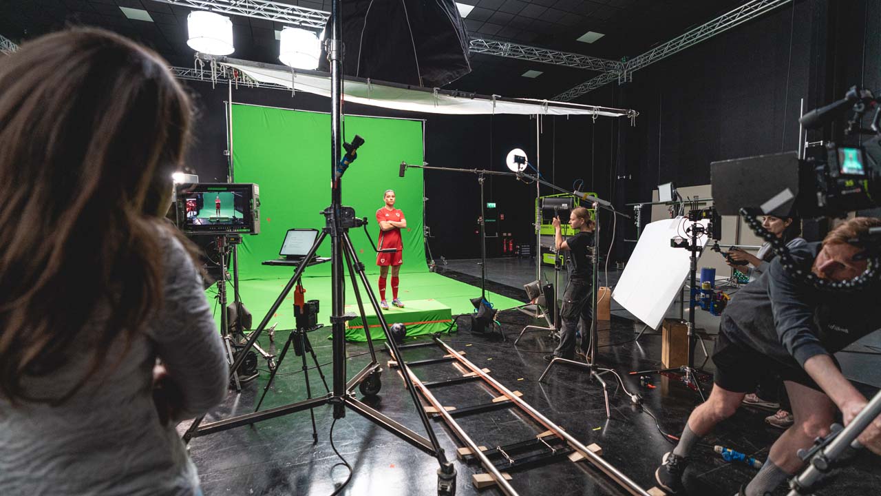 Film crew filming a woman in a red outfit standing in front of a green screen in a studio with lighting and camera equipment.