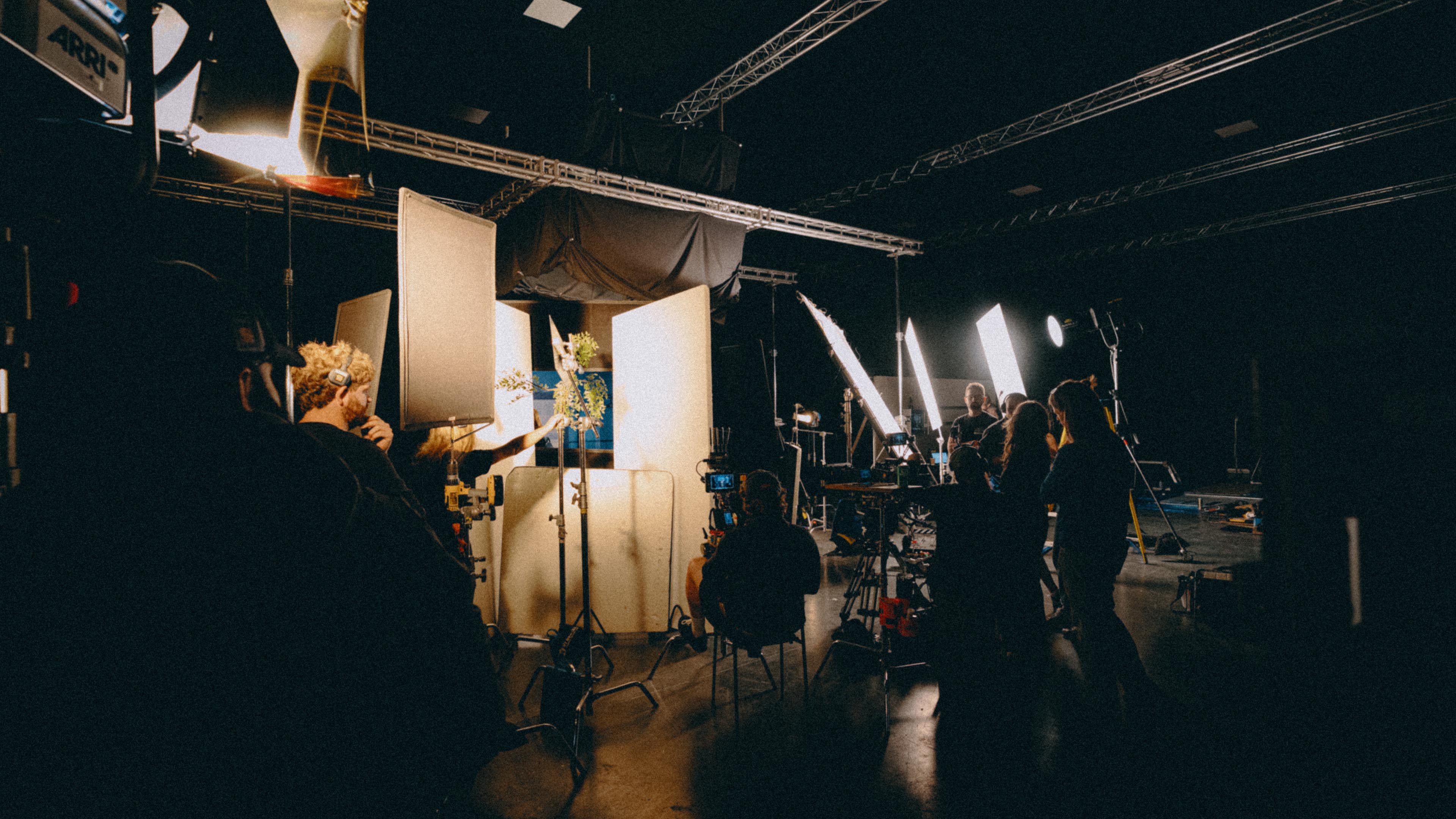 Film crew working on a dark set with professional lighting and cameras filming through white diffusion panels.