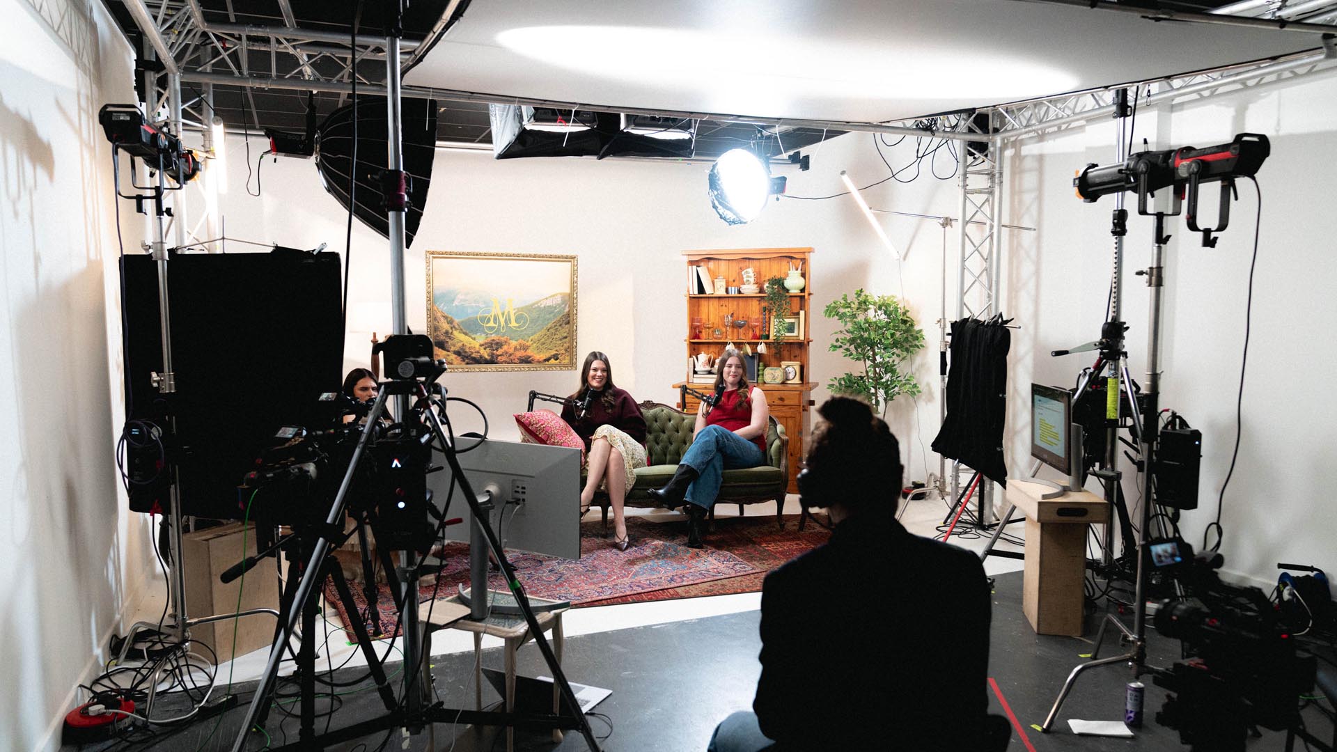 Two women sitting on a green sofa in a photography studio surrounded by lighting equipment and cameras.