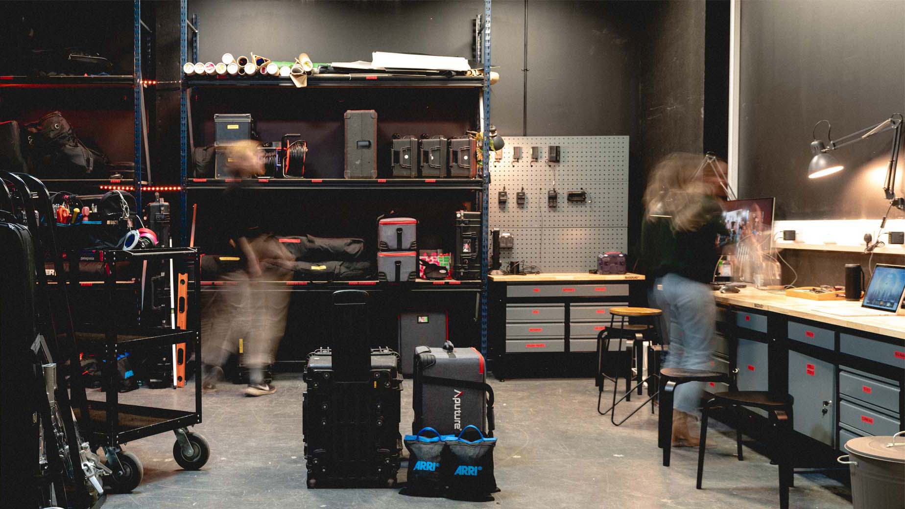 Studio equipment storage room with shelves, cases, gear, and two blurred figures moving inside.