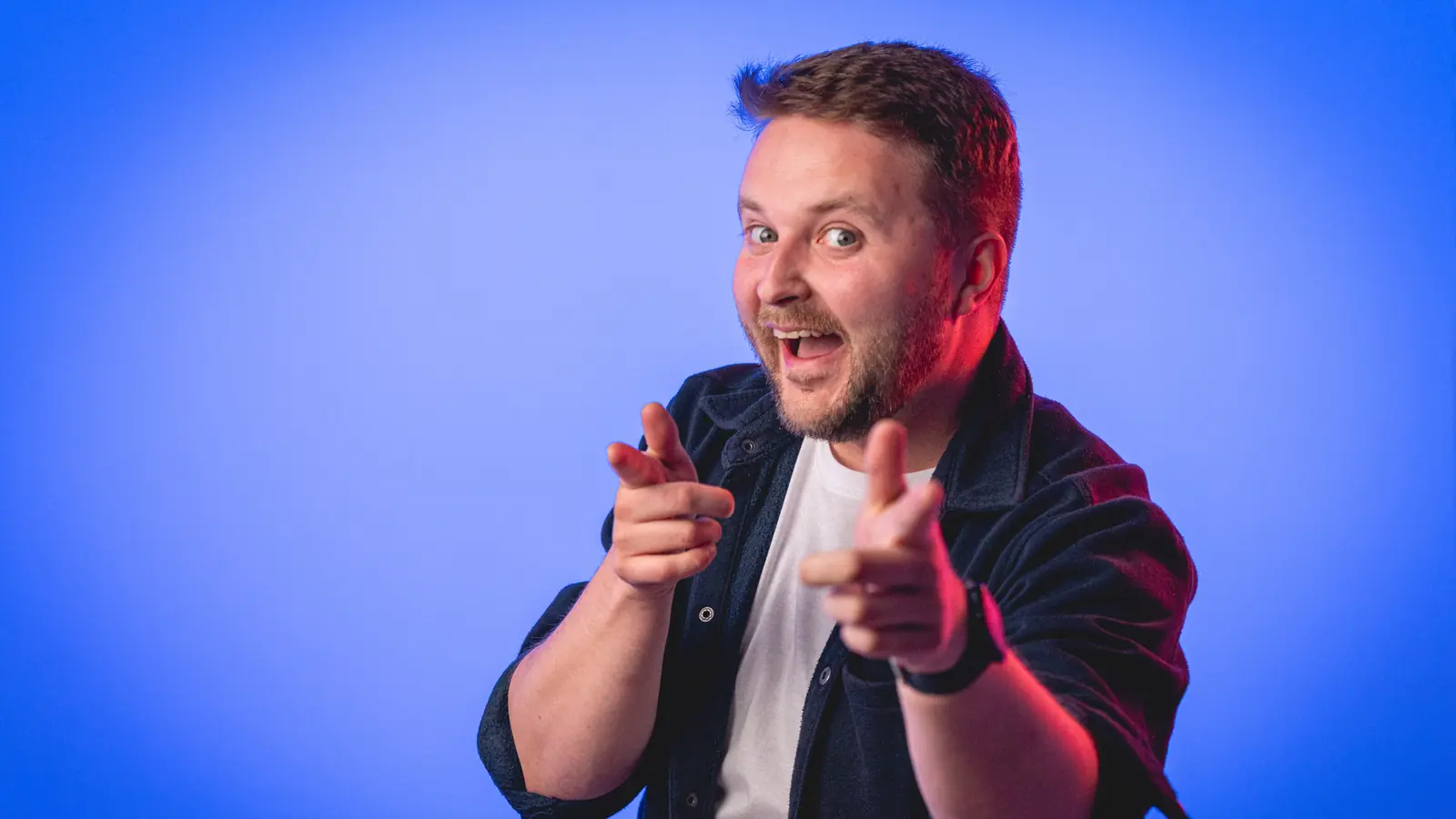 Smiling man with beard pointing finger guns towards the camera against a blue background with red lighting on the side.