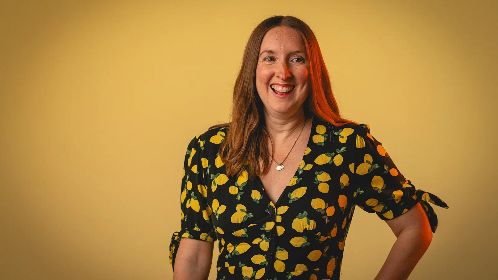 Smiling woman with long brown hair wearing a black dress with yellow lemon prints, standing against a yellow background.