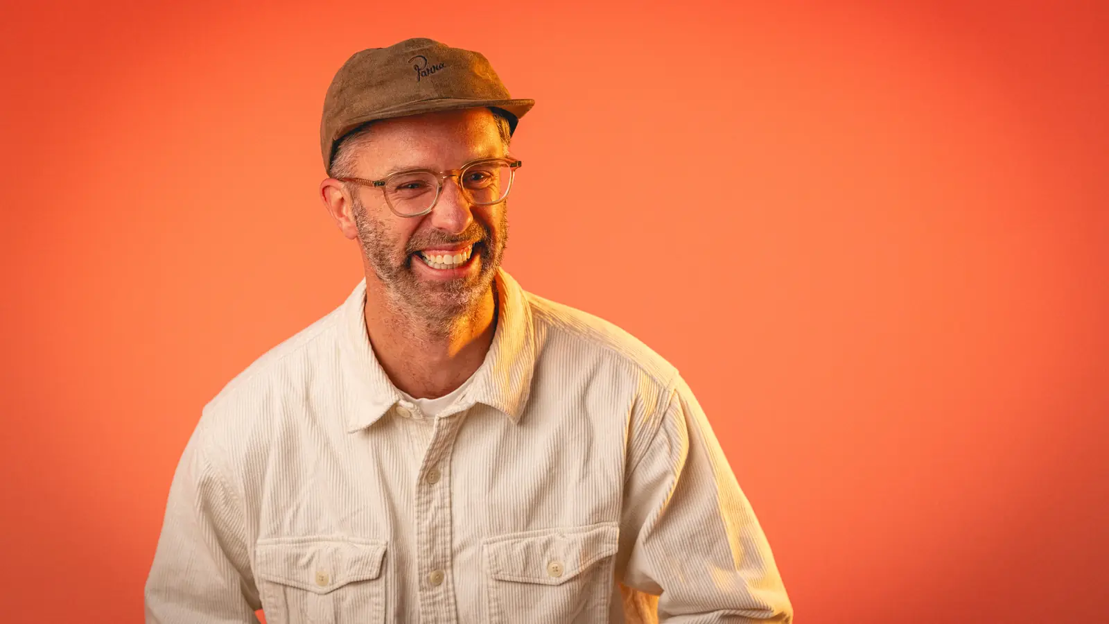Smiling man with glasses and a brown cap wearing a white shirt against an orange background.