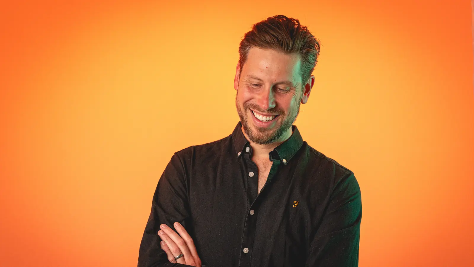 Smiling man with beard wearing a black button-up shirt standing with arms crossed against an orange background.