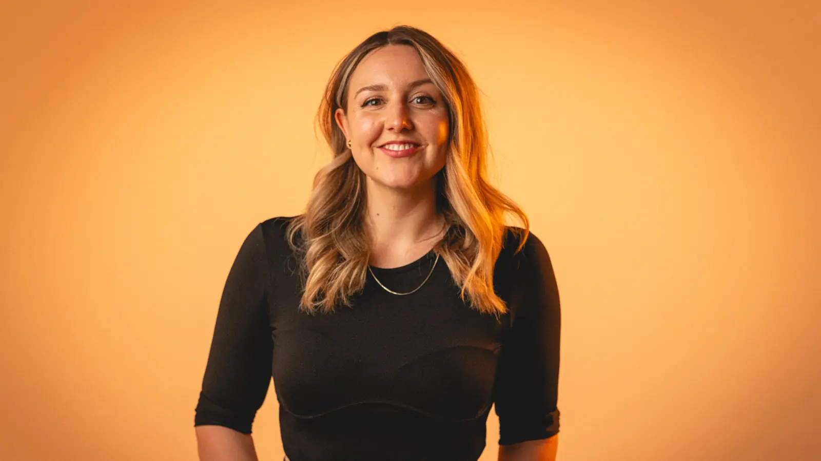 Smiling woman with wavy blonde hair wearing a black top against an orange background.