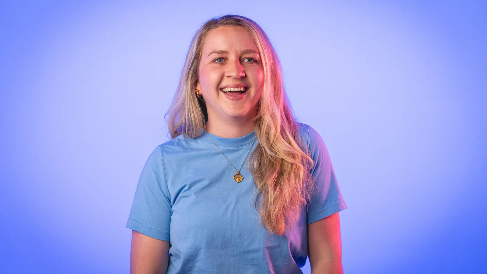 Smiling woman with long blonde hair wearing a light blue t-shirt and a gold pendant necklace against a blue and purple gradient background.