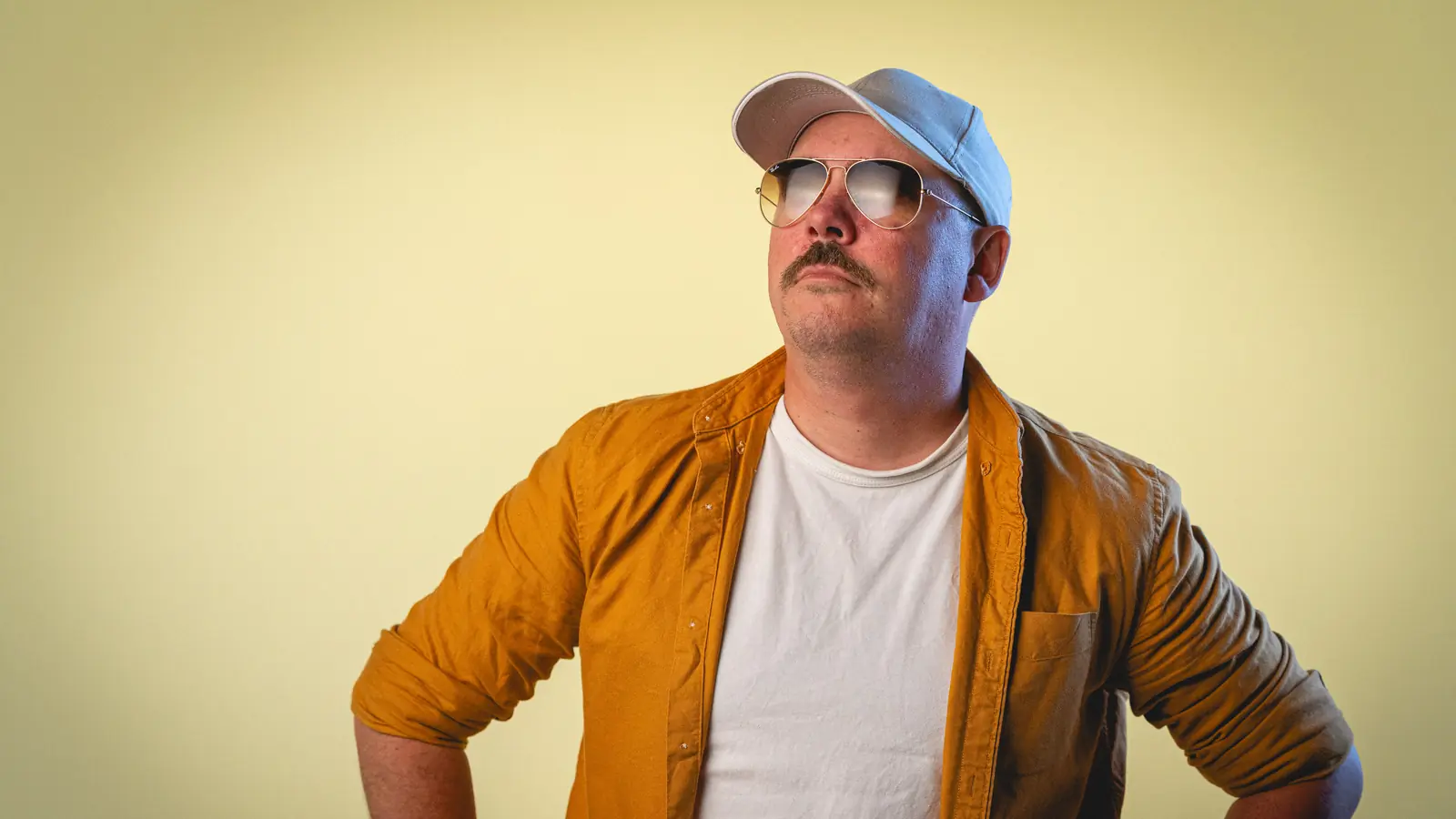 Man with mustache wearing a white cap, aviator sunglasses, a white shirt, and a mustard yellow jacket, posing confidently with hands on hips against a yellow background.
