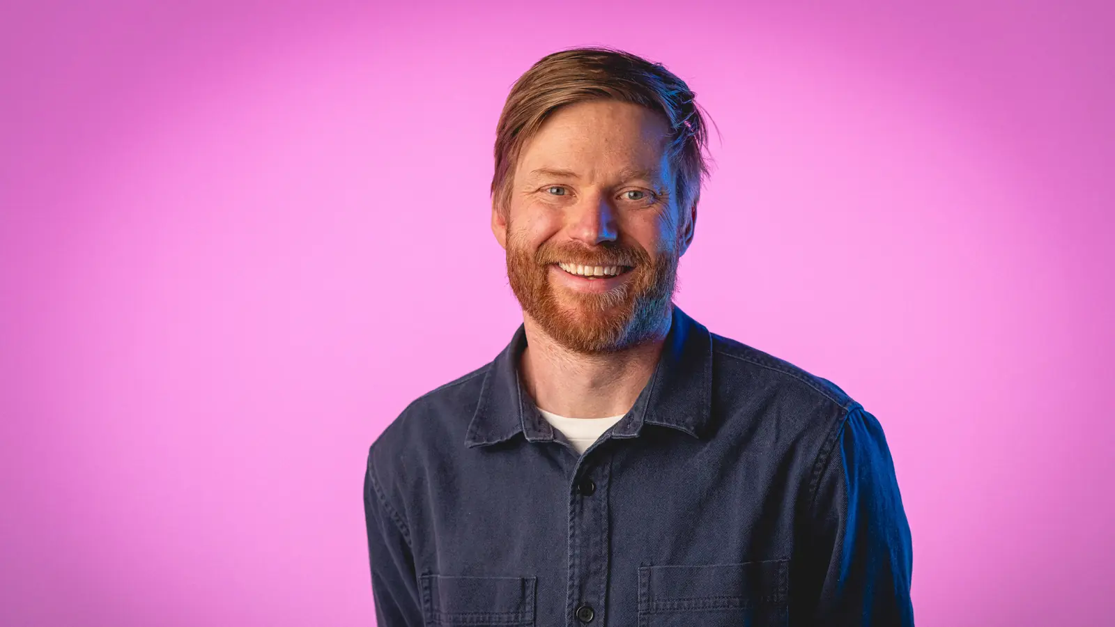Smiling man with light brown hair and beard wearing a dark button-up shirt against a pink background.