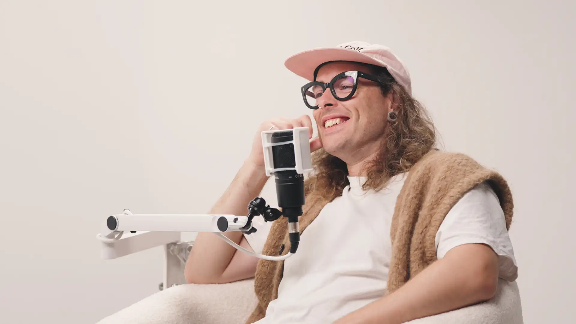 Smiling man with long hair wearing glasses and a cap sitting in a chair speaking into a microphone on a white arm stand.