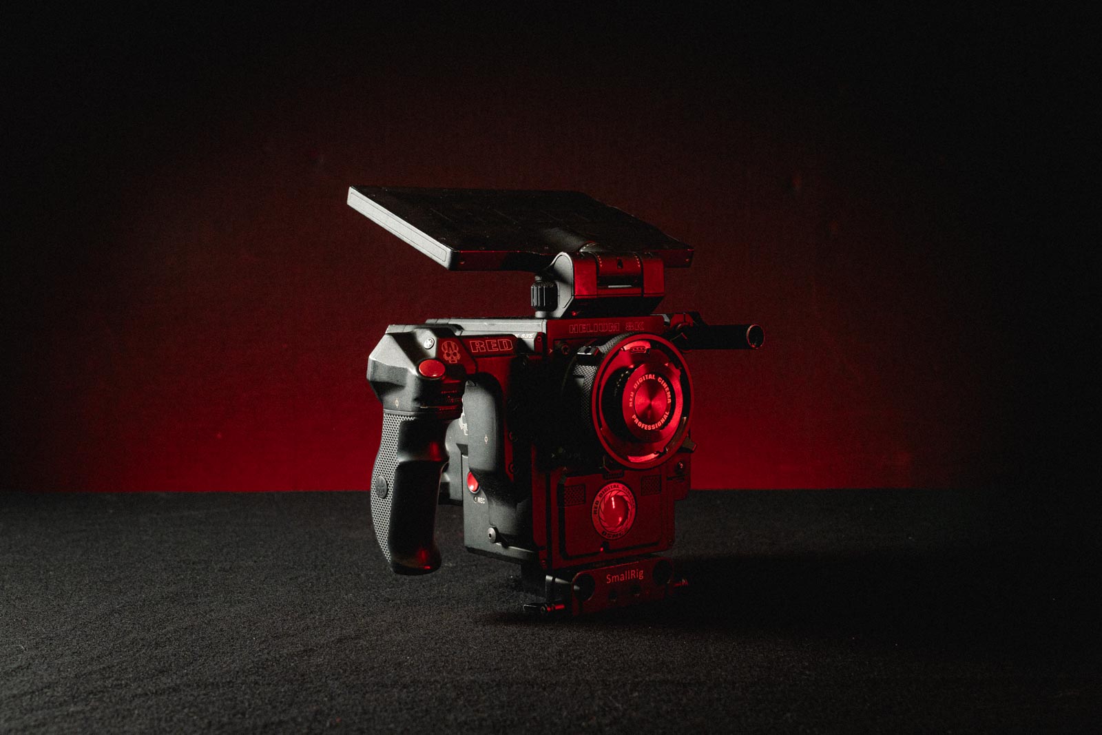 Professional RED digital cinema camera with mounted monitor and grip handles on a dark surface with red backlighting.