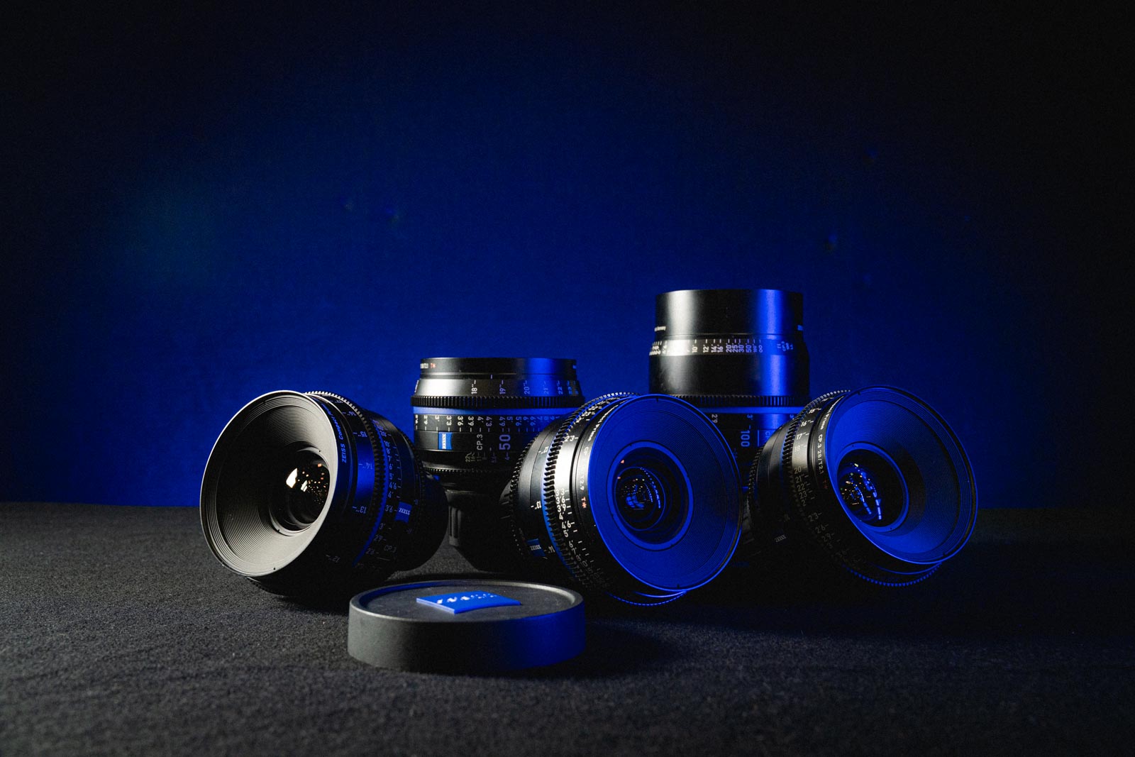 Four professional camera lenses arranged on a black surface with blue lighting and a lens cap in front.