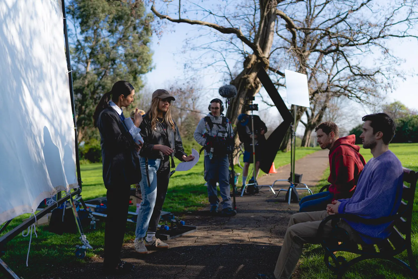 Film crew with boom mic and camera filming two men sitting on a park bench during a daytime shoot.