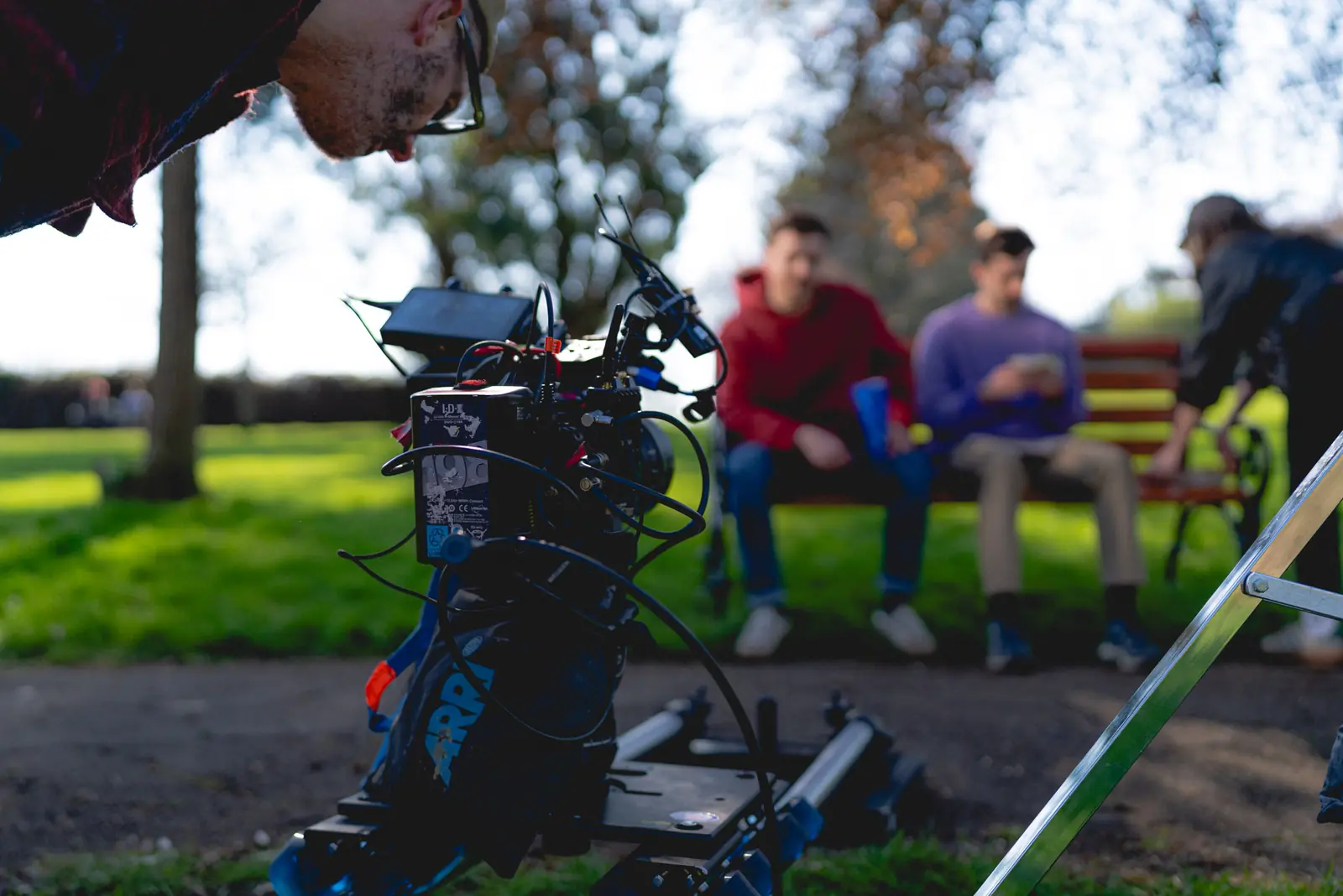 Man operating professional film camera on a dolly in a park while two people sit on a bench in the background.