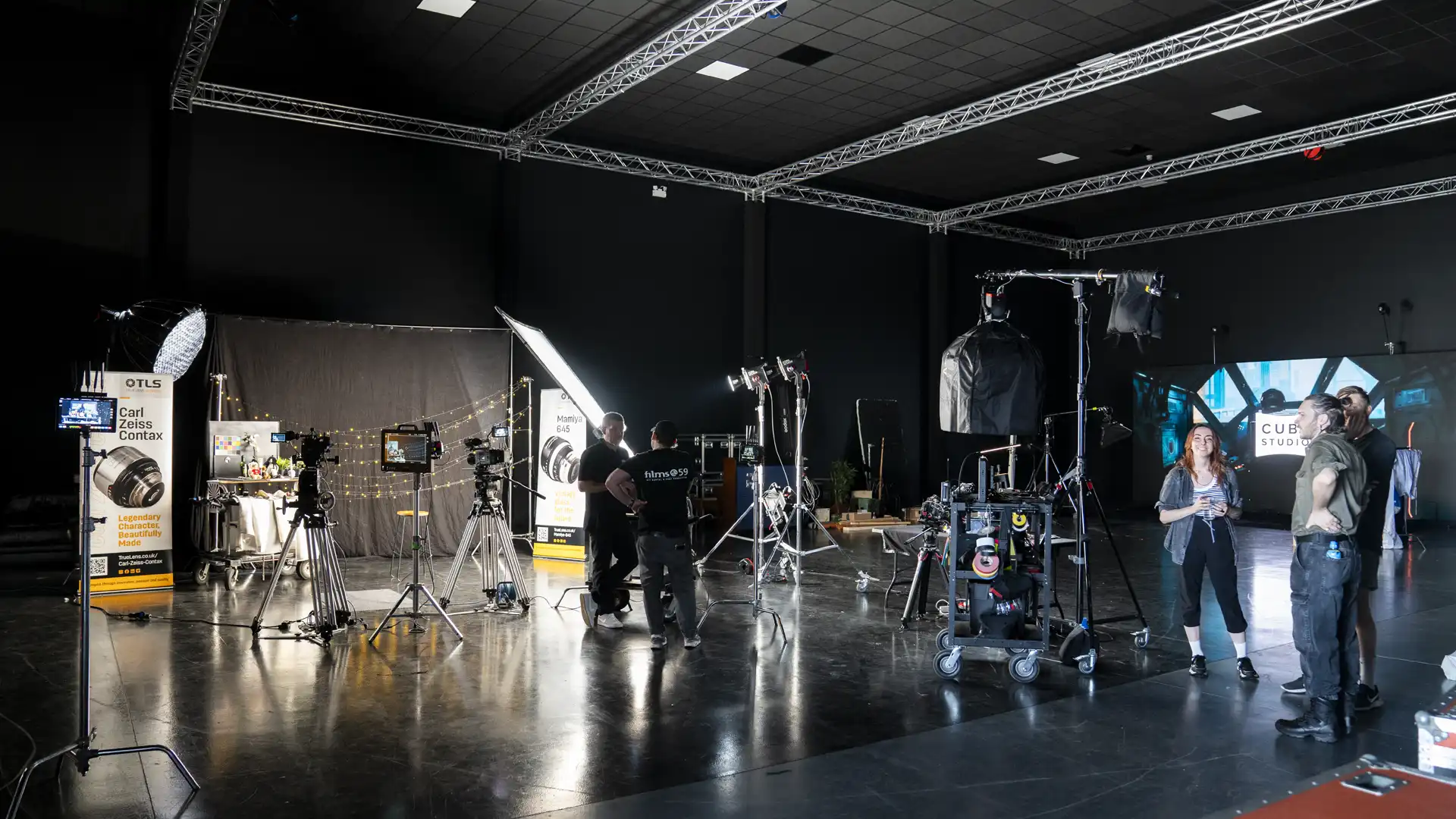 Film studio set with professional lighting equipment, cameras on tripods, and crew members discussing and preparing for a shoot against a dark backdrop.