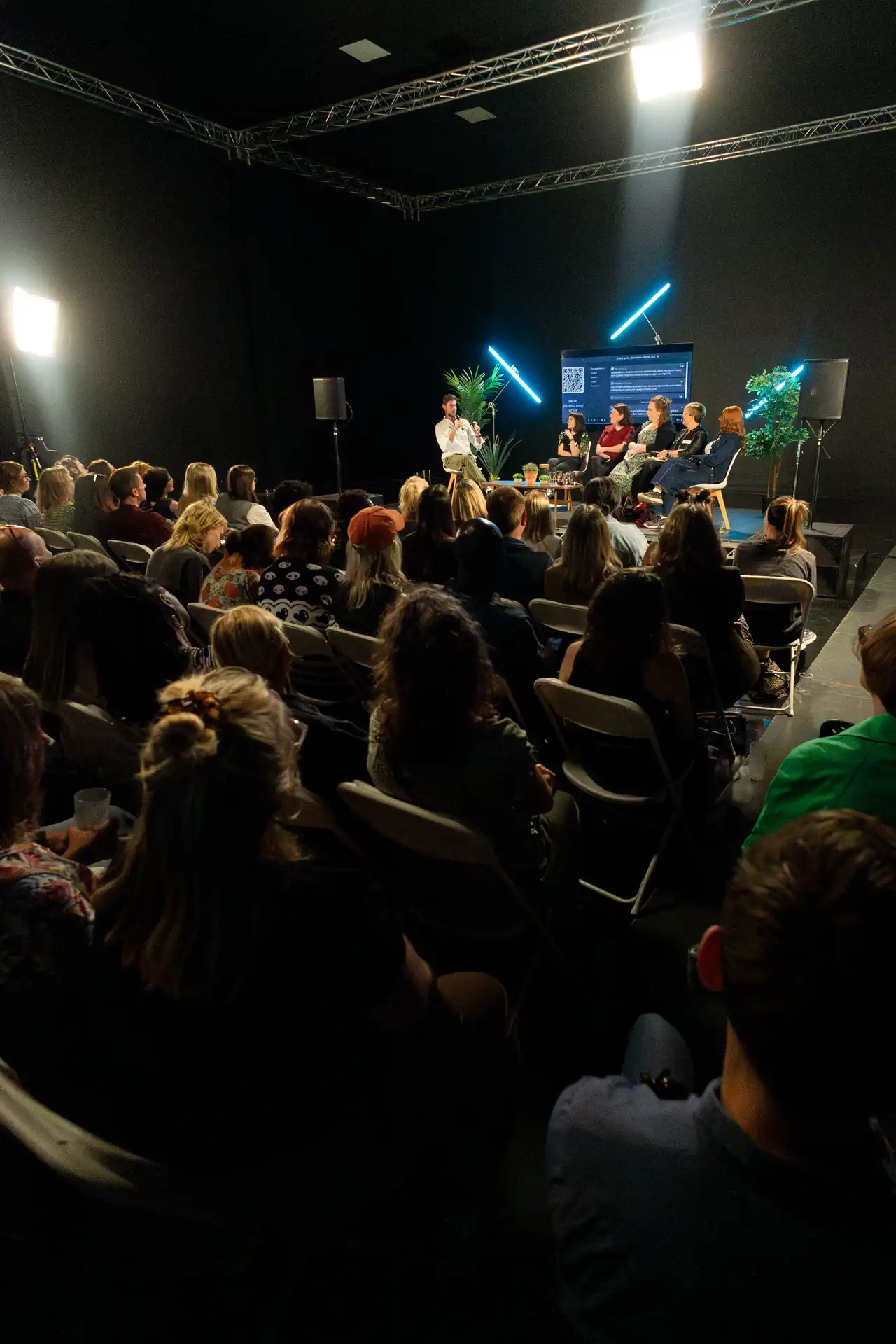 Audience listening to a panel discussion of five people on stage with a screen and decorative plants in a dark room.
