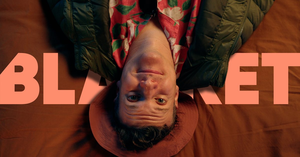 Man with floral shirt, green jacket, and red hat lying upside down against a brown background with the word 'BLANKET' in large orange letters.