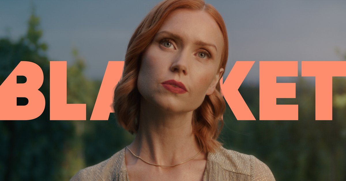 Portrait of a red-haired woman with light skin and neutral expression wearing a gold necklace and top, with large orange text partially visible behind her.