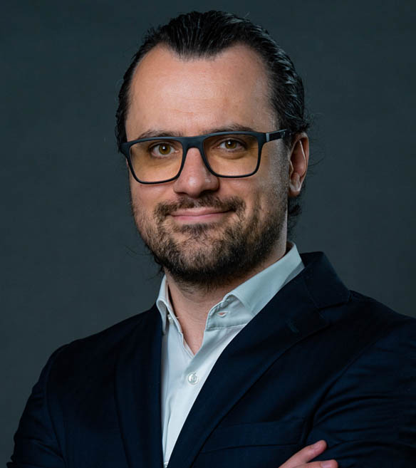 Bearded man wearing glasses, dark jacket, light shirt, against a dark gray background