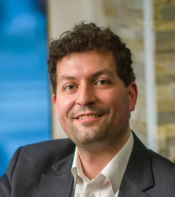 A bearded, curly-haired, smiling man in a suit against a blue-gray background