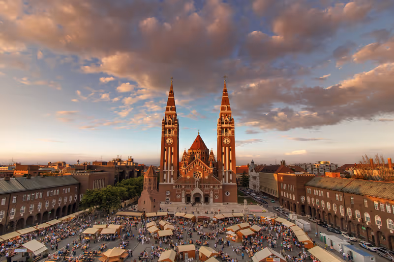The imposing facade and two tall towers of Szeged Cathedral in the evening light, with a square crowded with tents and people in front of it.