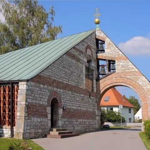 A small stone church with a green metal roof and a bell tower with three bells, behind it a blue sky and a white house with a red roof.