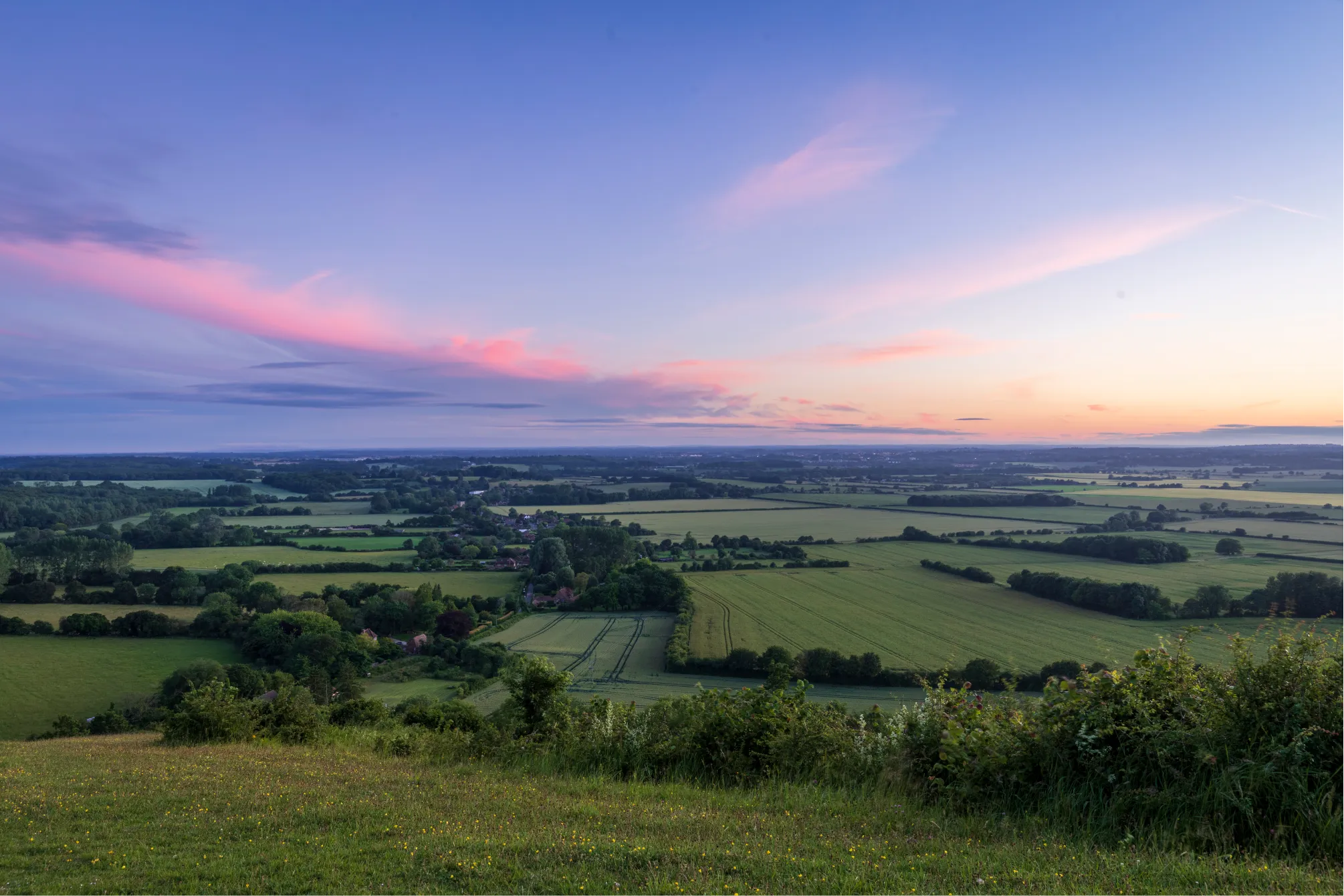 Expansive rural landscape with green fields, hedgerows, and a pink-tinged sunset sky.
