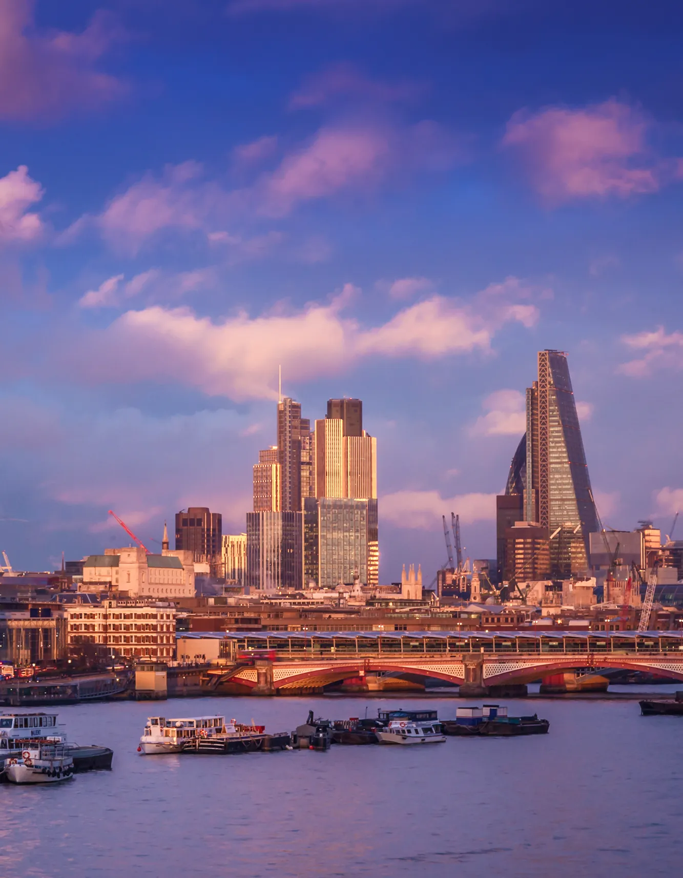 View of London's city skyline at sunset with boats on the river and a bridge in the foreground.
