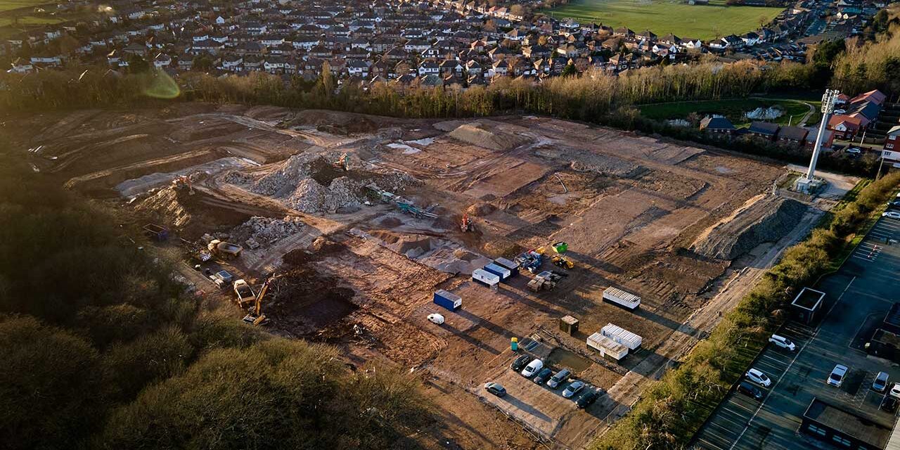 Aerial view of a large brownfield construction site with earthworks, machinery, and temporary buildings near a residential area.