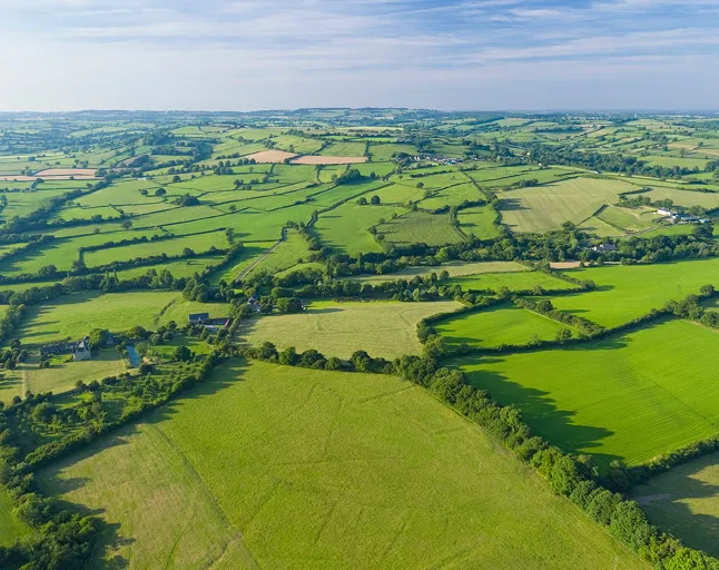 Aerial view of a patchwork of green farmland fields separated by hedgerows under a partly cloudy sky.