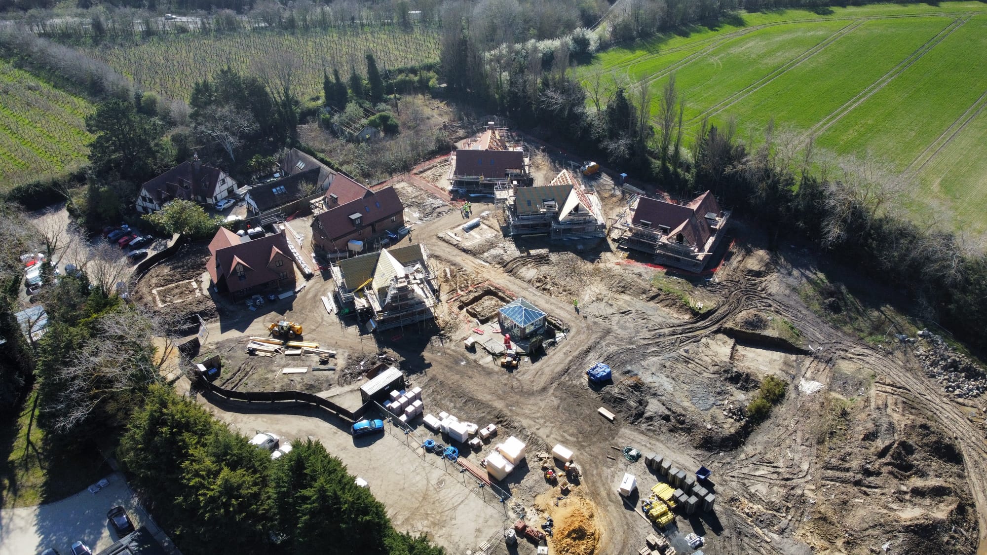 Aerial view of a residential construction site with several houses in various stages of building surrounded by trees and green fields.