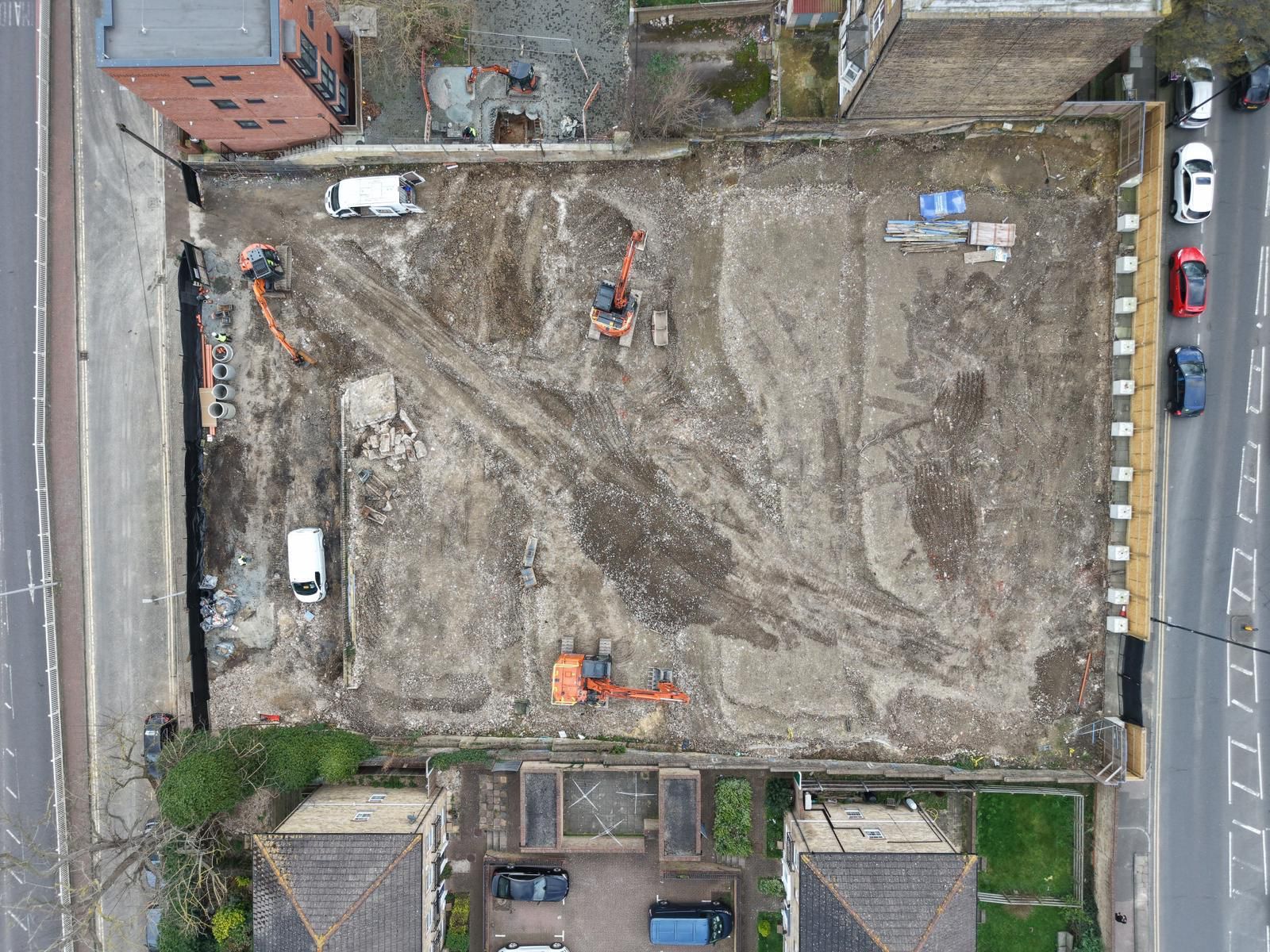 Aerial view of a construction site with excavators, trucks, dirt, and two bordering roads with parked cars.