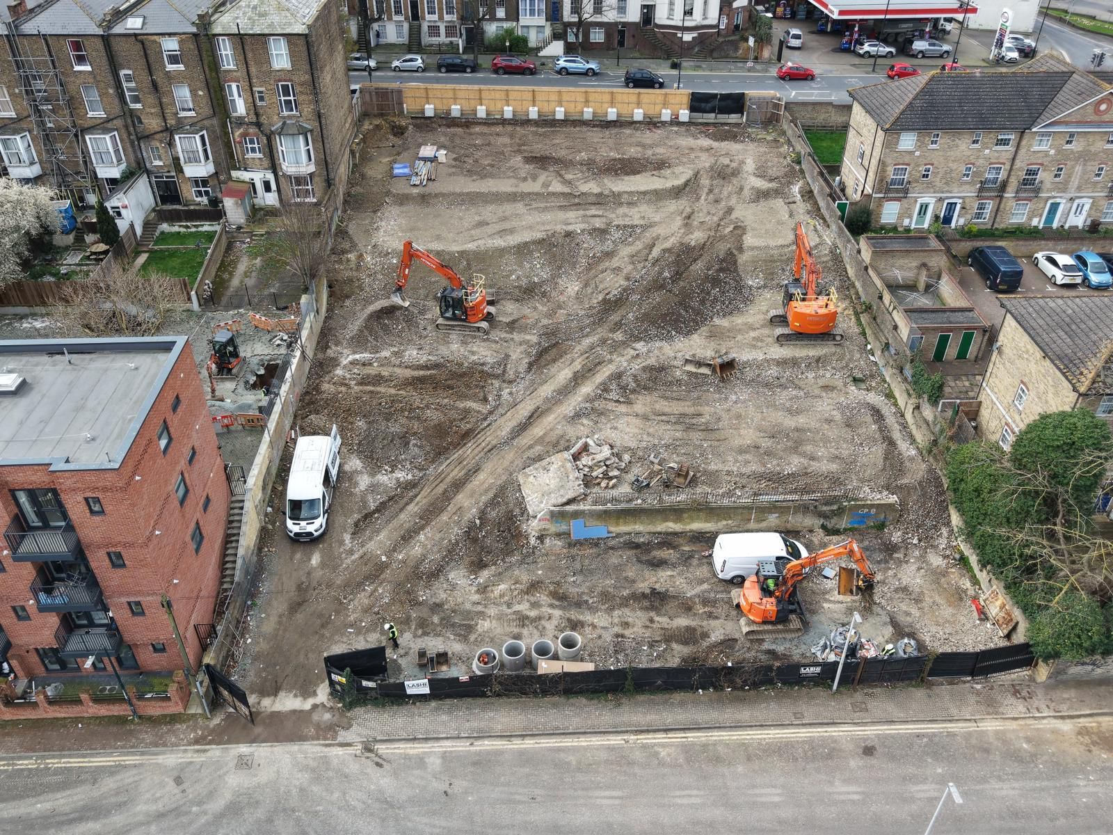 Aerial view of a construction site with three orange excavators, a white van, and several concrete pipes surrounded by residential buildings and a street.