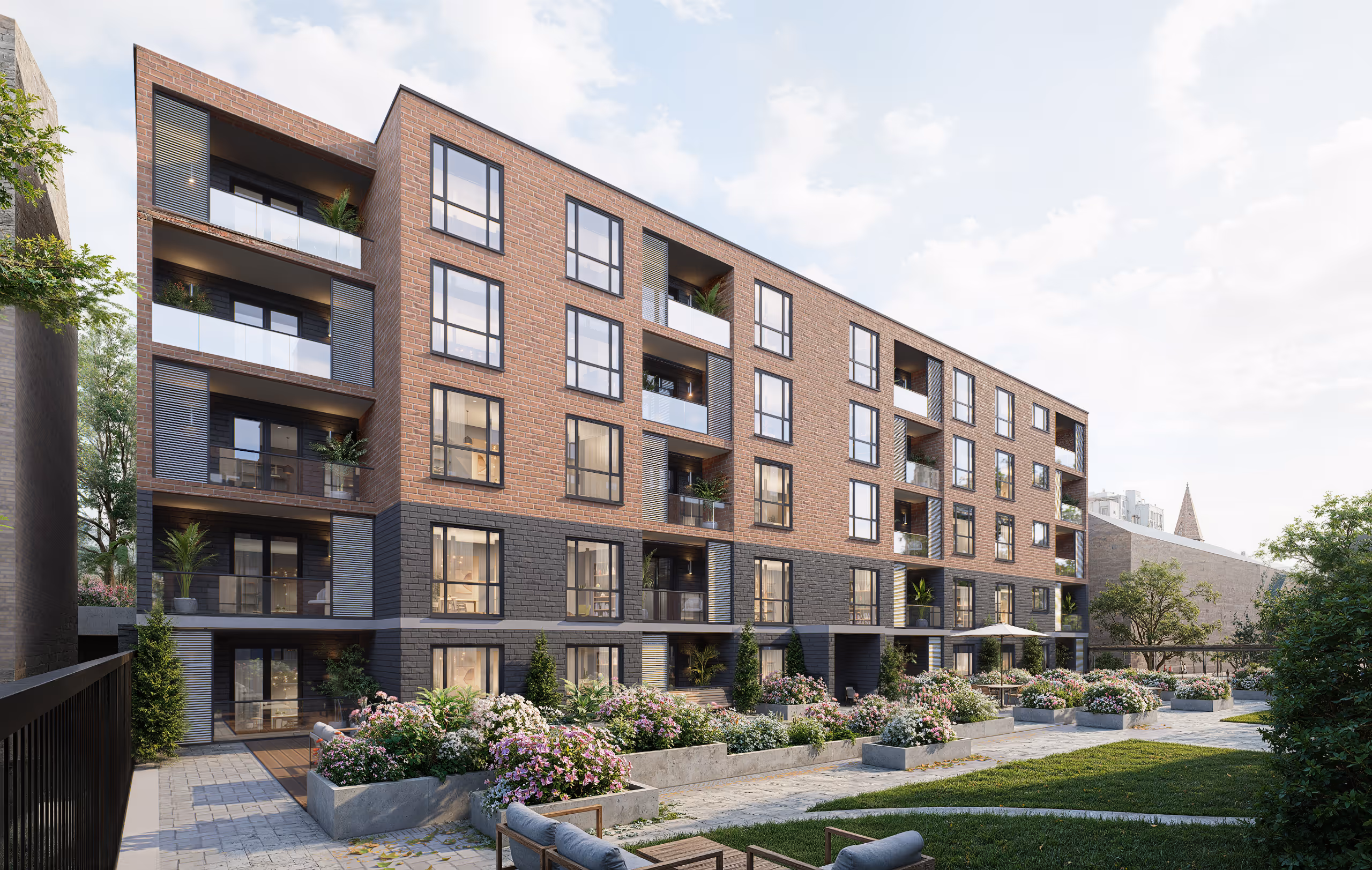 Modern brick apartment building with balconies overlooking landscaped garden beds and outdoor seating area under a partly cloudy sky.