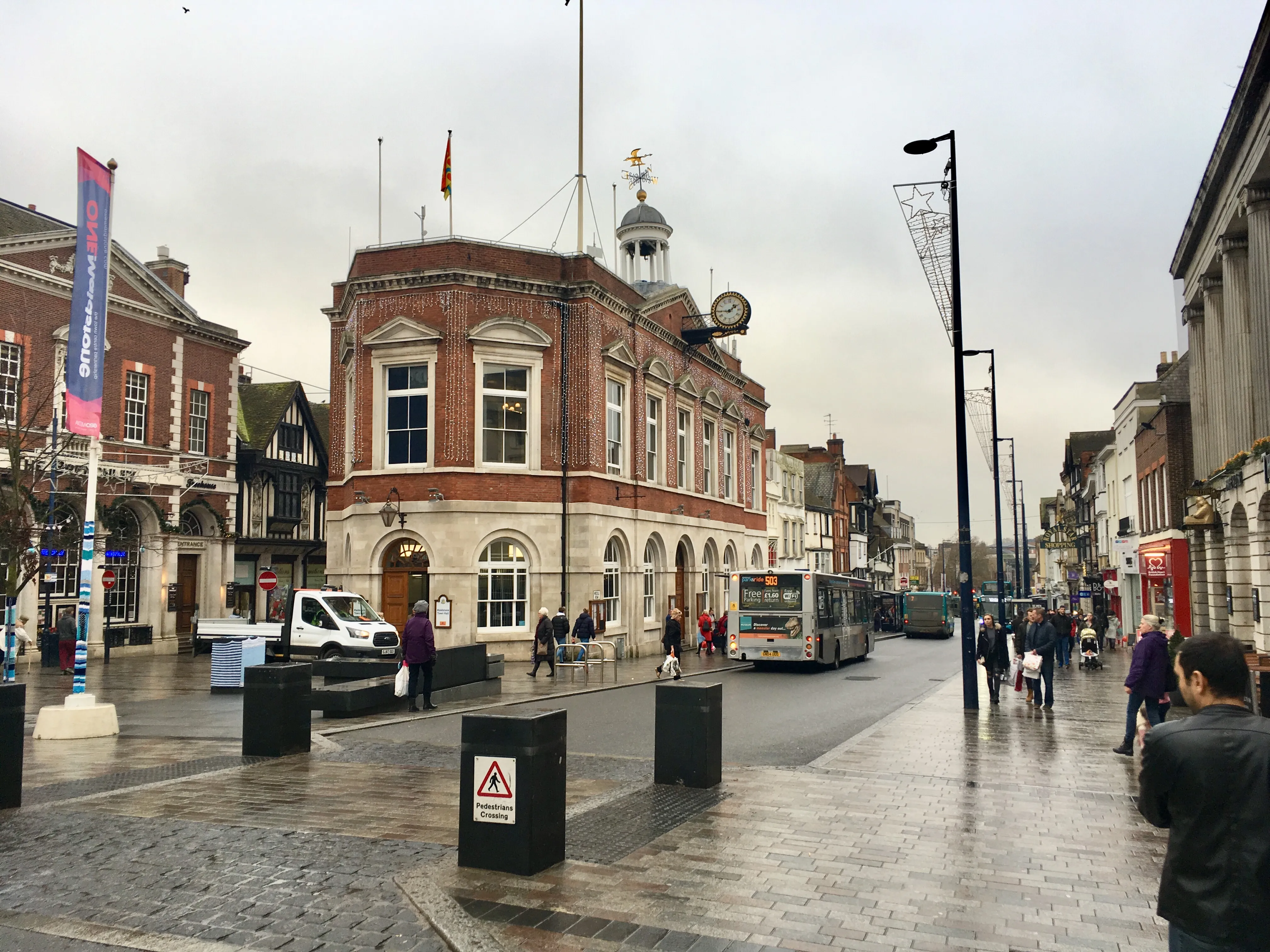 Wet street scene on a cloudy day in a town with historic brick buildings, pedestrians walking, and buses on the road.