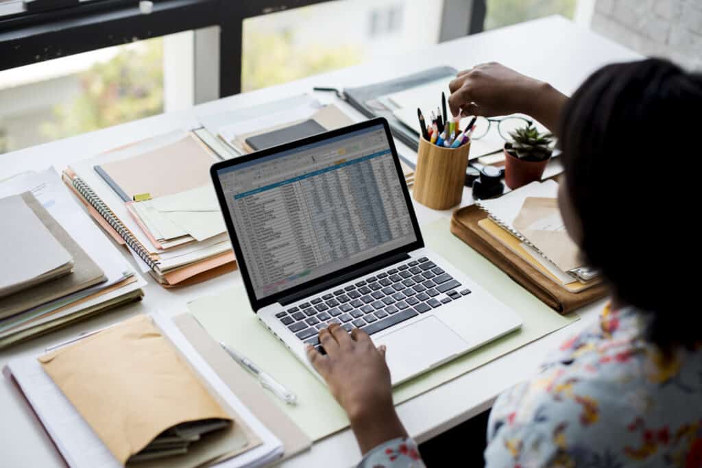 Person working on laptop at desk with notebooks, supplies, and small plant.