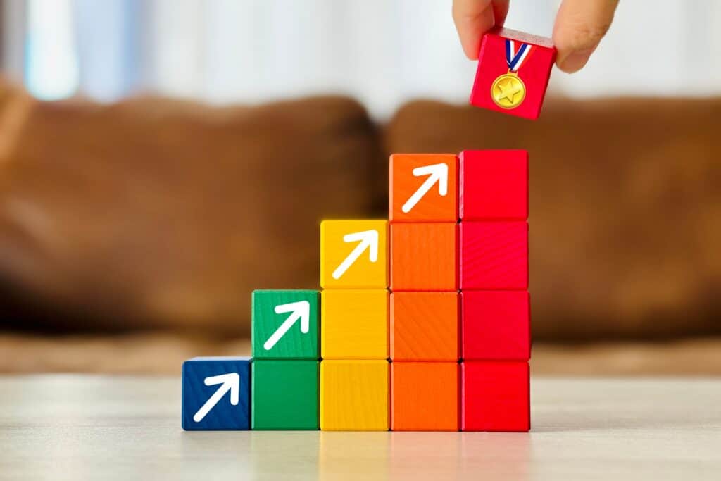 Person placing gold medal on stacked colorful blocks with upward arrows symbolizing growth and success.