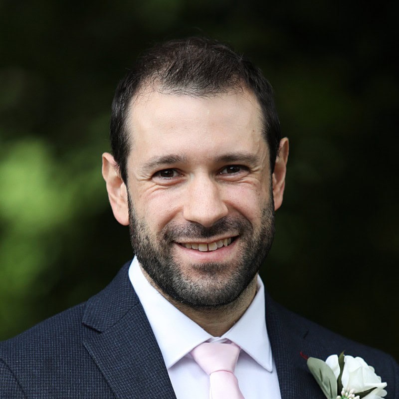 Smiling man in dark suit with pink tie and white flower boutonnière outdoors
