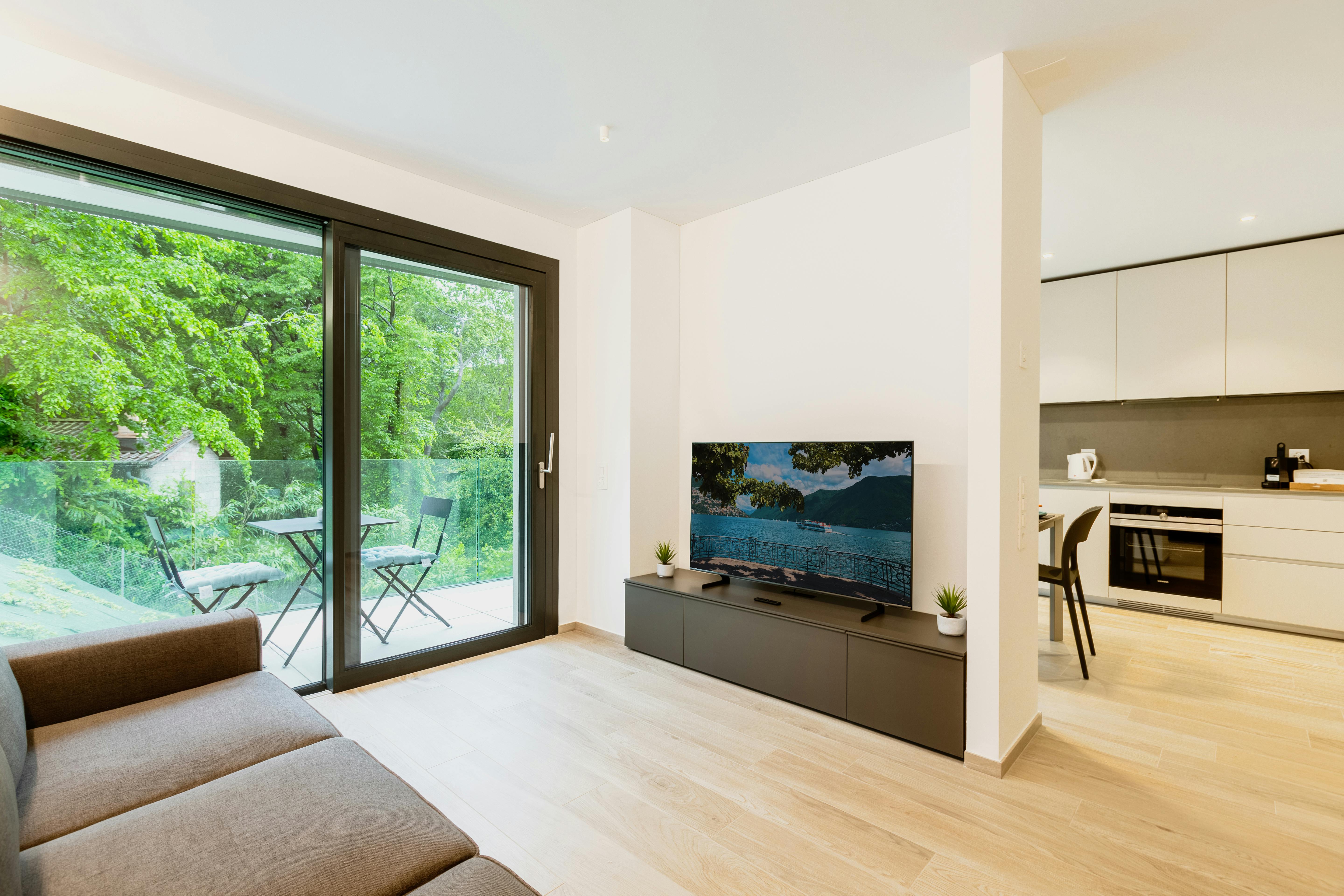Moderner openspace Wohnbereich mit grauem Sofa, großem Fenster mit Blick auf Terrasse, sowie angrenzender offener Küche.