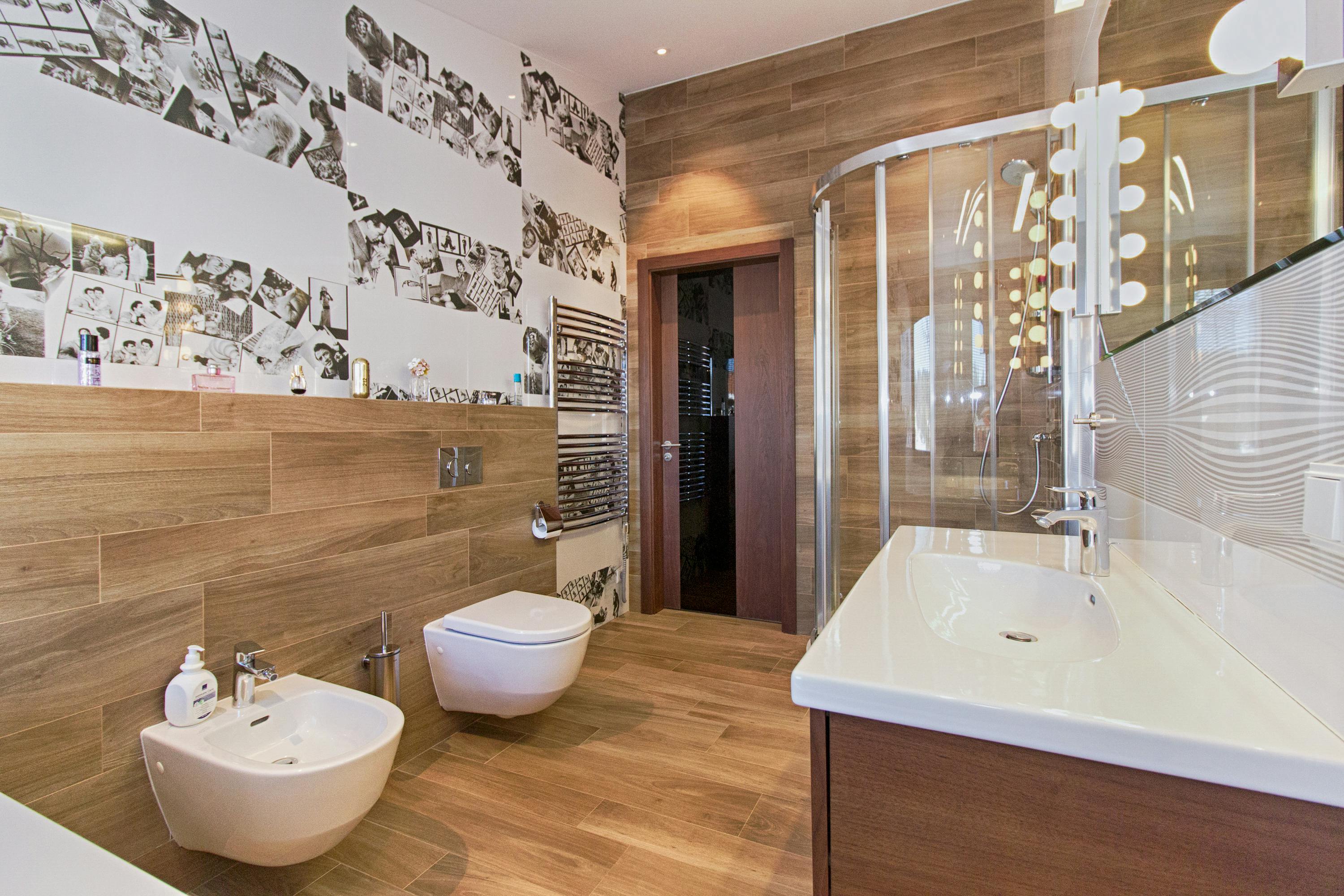 Großes modernes Badezimmer mit Holzboden, hängendem WC, Bidet, Glasdusche und Wand mit Schwarz-Weiß-Fotocollage.   