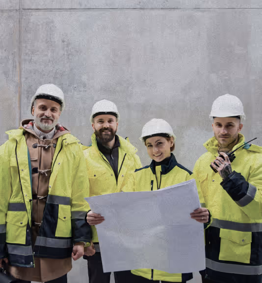 Four construction workers wearing white helmets and yellow reflective jackets reviewing a blueprint against a plain gray wall.