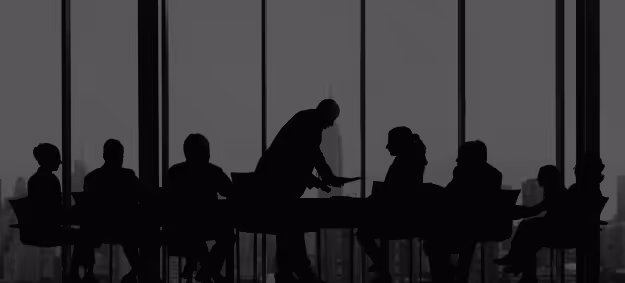 Silhouettes of a group of people seated around a conference table with one person standing, engaging with others in a modern office with large windows.