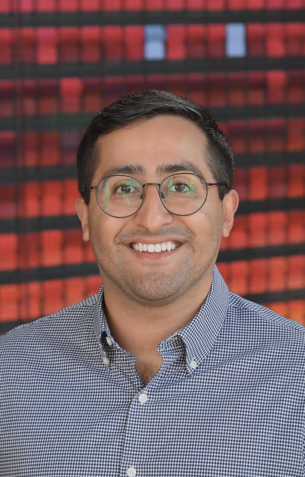 Smiling man with short dark hair, wearing round glasses and a checkered button-up shirt, with a blurred red and black background.