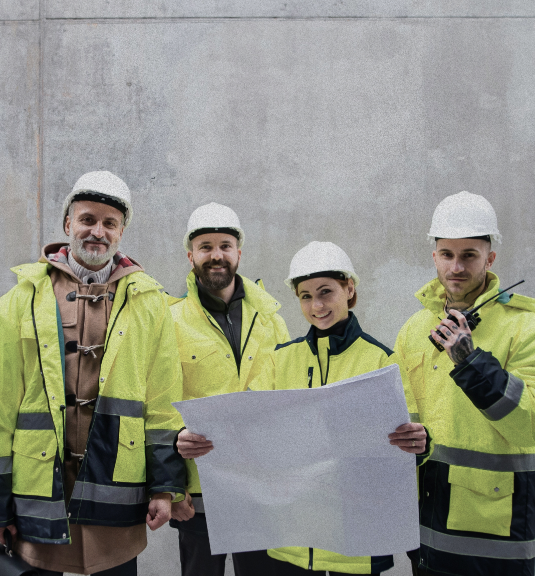 Four construction workers wearing white helmets and yellow reflective jackets reviewing a blueprint against a plain gray wall.