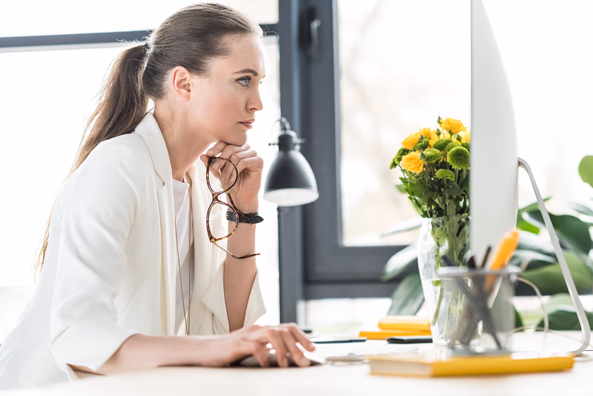 Frau im hellen Blazer arbeitet konzentriert am Bildschirm, Blumen und Pflanze daneben