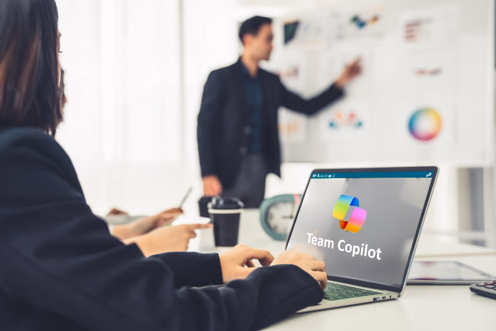 Frau am Laptop mit Team-Copilot-Logo auf dem Bildschirm, im Hintergrund Kollege an der Wandtafel